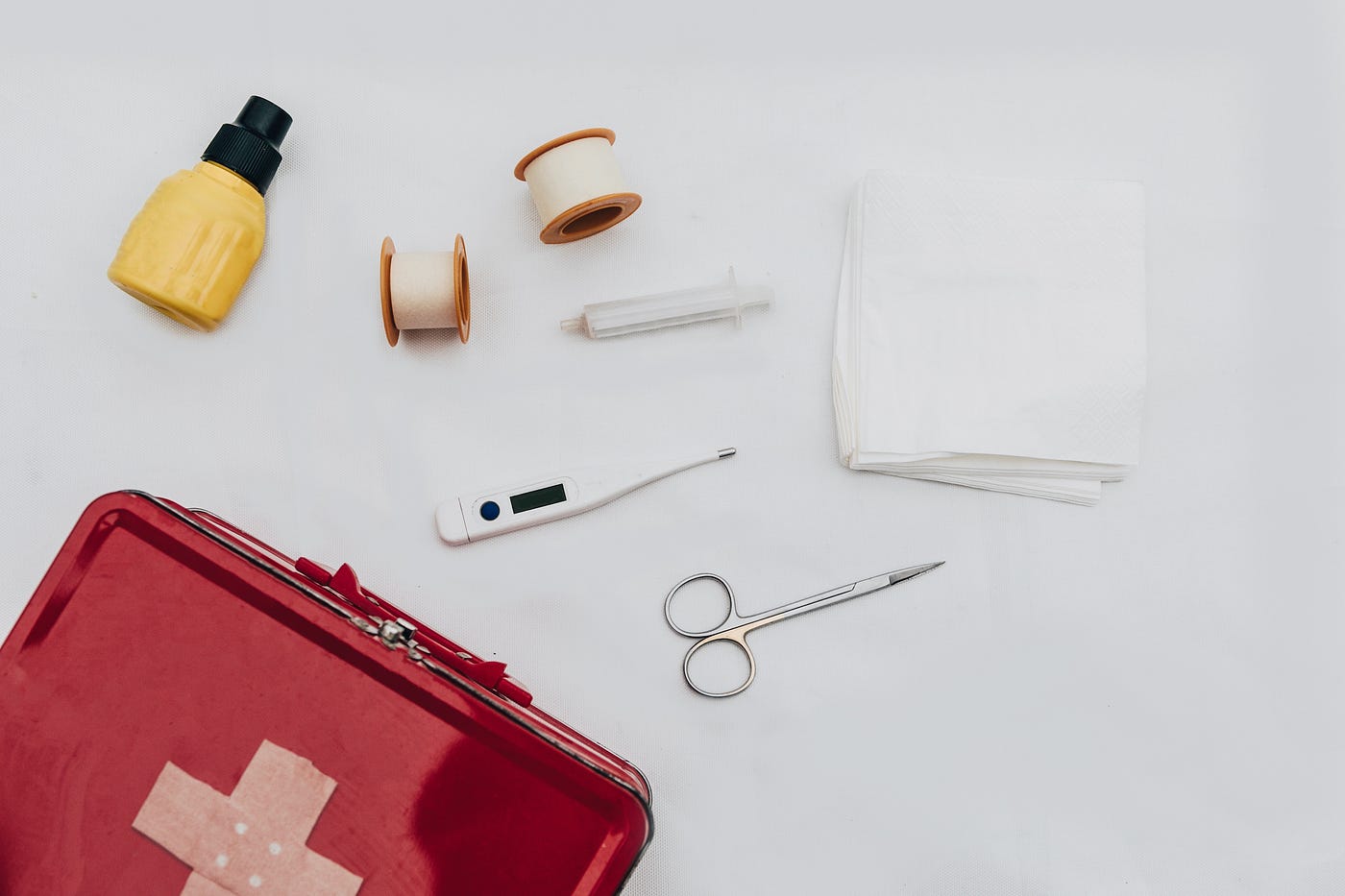 Overhead view of a red first aid kit and some of its contents: thermometer, scissors, gauze, and a small yellow bottle.