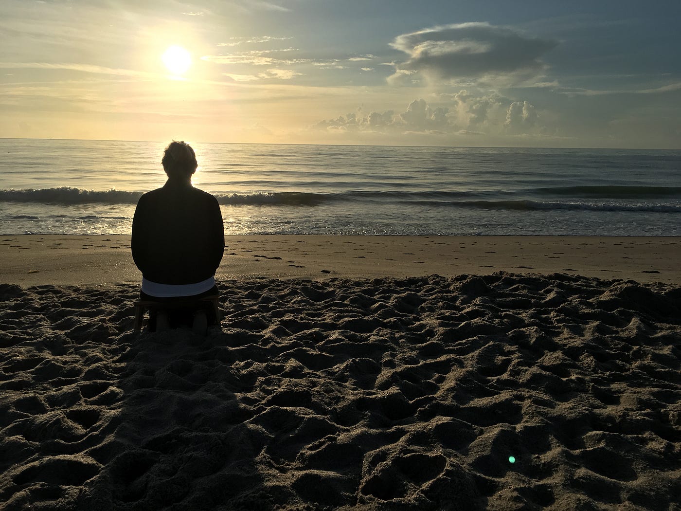 해변에 앉아 일출을 바라보며 명상하는 한 여성의 뒷모습.