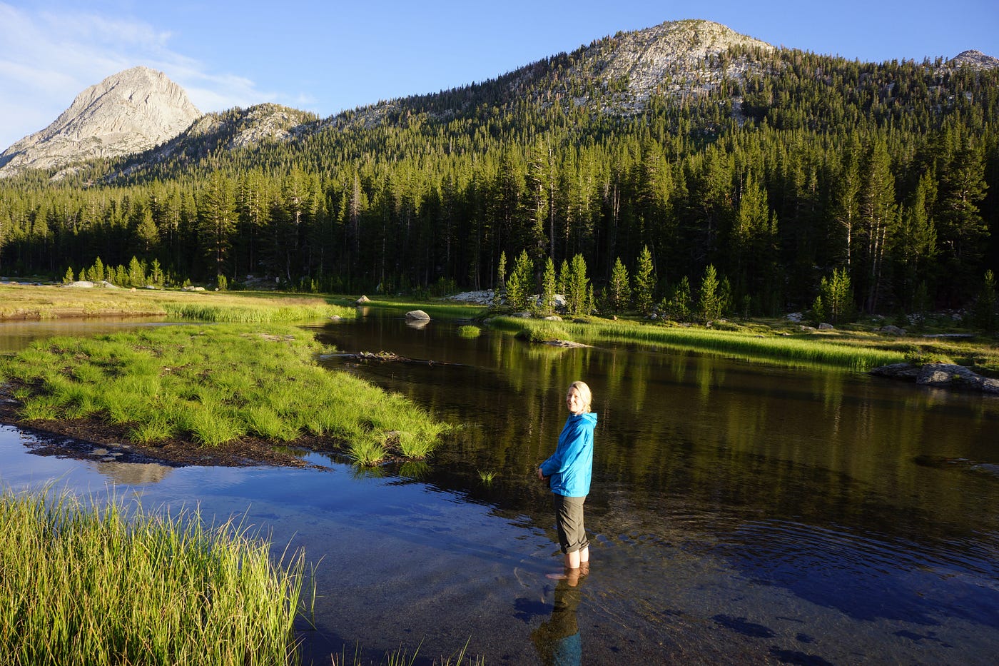 A person standing in a river with forested mountains in the background on a clear day.