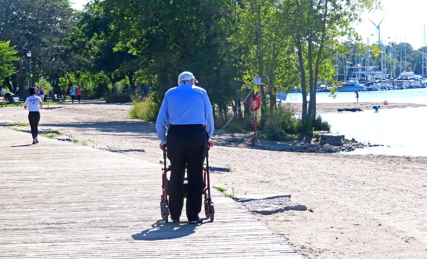 나이 든 남자가 나무 판자길에서 보행기를 사용하고, 그 앞에는 조깅하는 사람이 있고, 오른쪽에는 해변이 보이고, 조금 더 앞에는 나무가 서 있습니다.<BR>