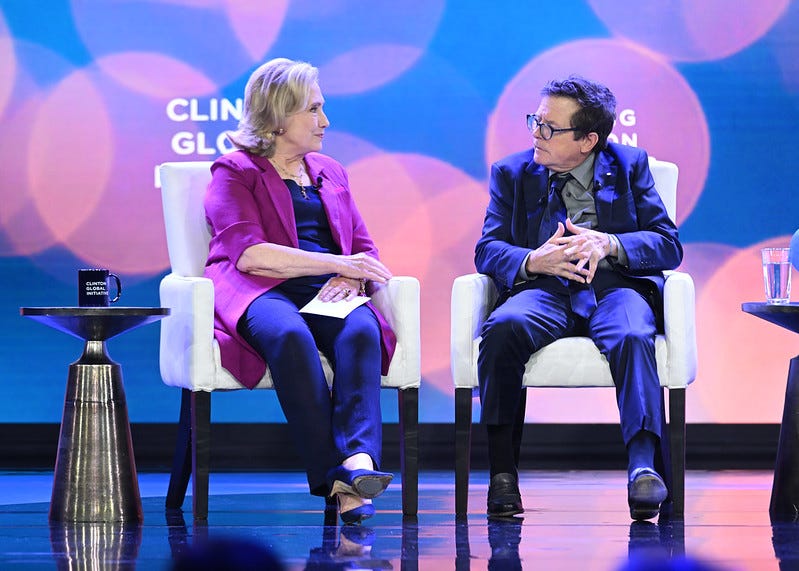 Secretary Clinton and Michael J. Fox sit in two chairs behind a blue background. They are facing each other and talking.