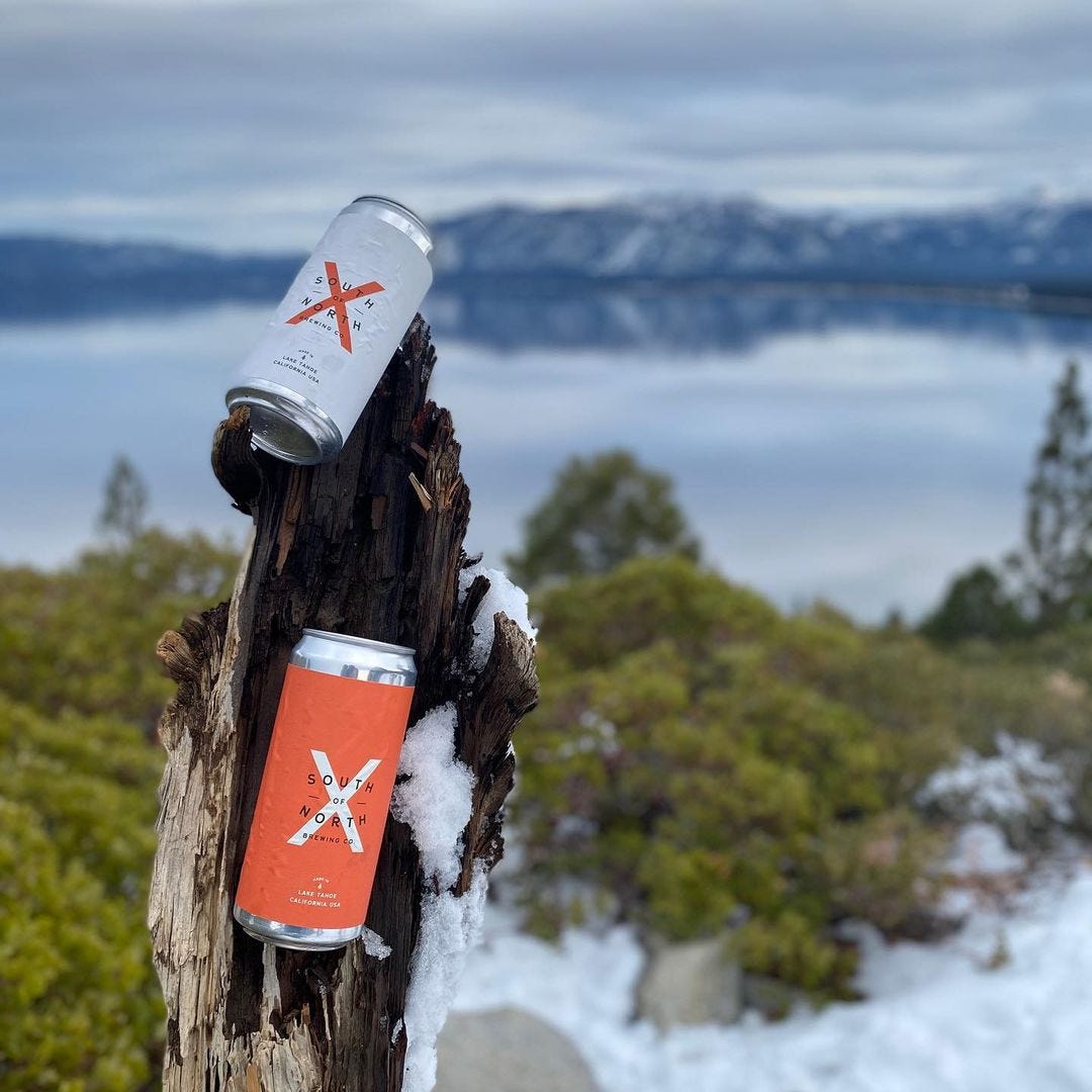 2 cans of beer with X’s on them perched on a stump with Lake Tahoe in the background.