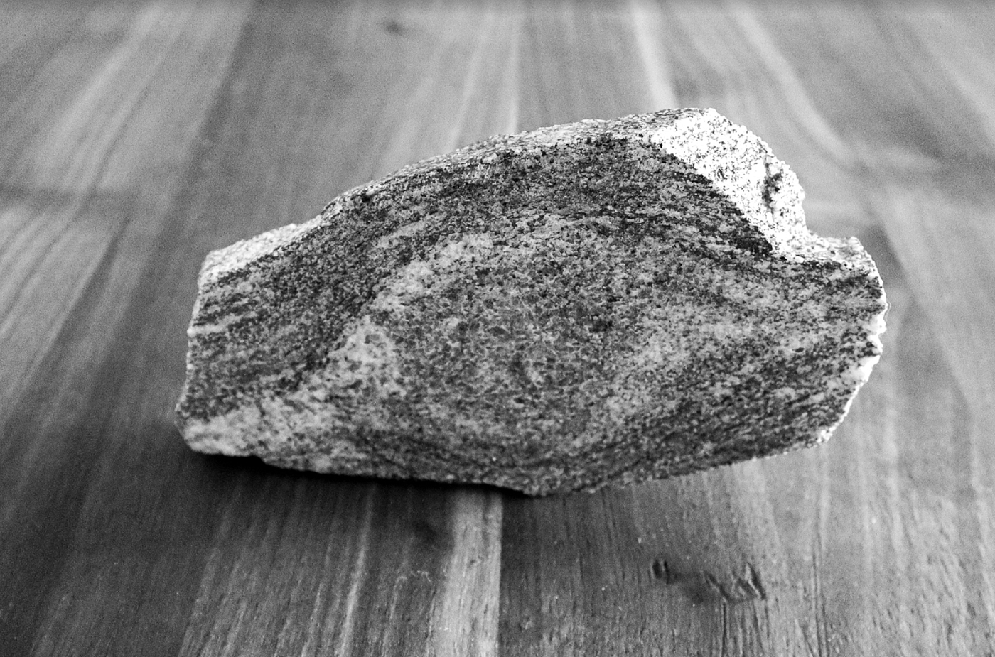 Photo: Francine Fallara: Pressure’s Beauty: A close-up black and white photograph of a metamorphic rock sample featuring a prominent, eye-shaped augen texture. The banded mineral layers curve gracefully around a dense central core. The specimen rests on a smooth wooden surface with linear grain patterns that run perpendicular to the rock’s internal structures.