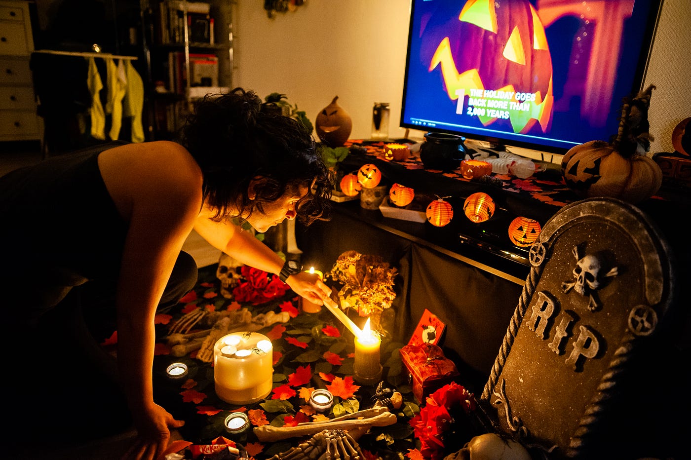 A person lights a candle on an altar on the floor in front of a TV displaying a jack-o’-lantern.
