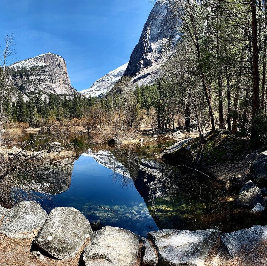 Two snowy mountains reflected in a perfectly still body of water ringed by trees.