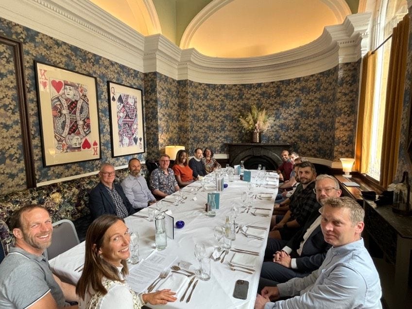 Tech leaders are sat round a long rectangular table in a historic room. All smiling and looking at the cammara. Ready for lunch to begin.