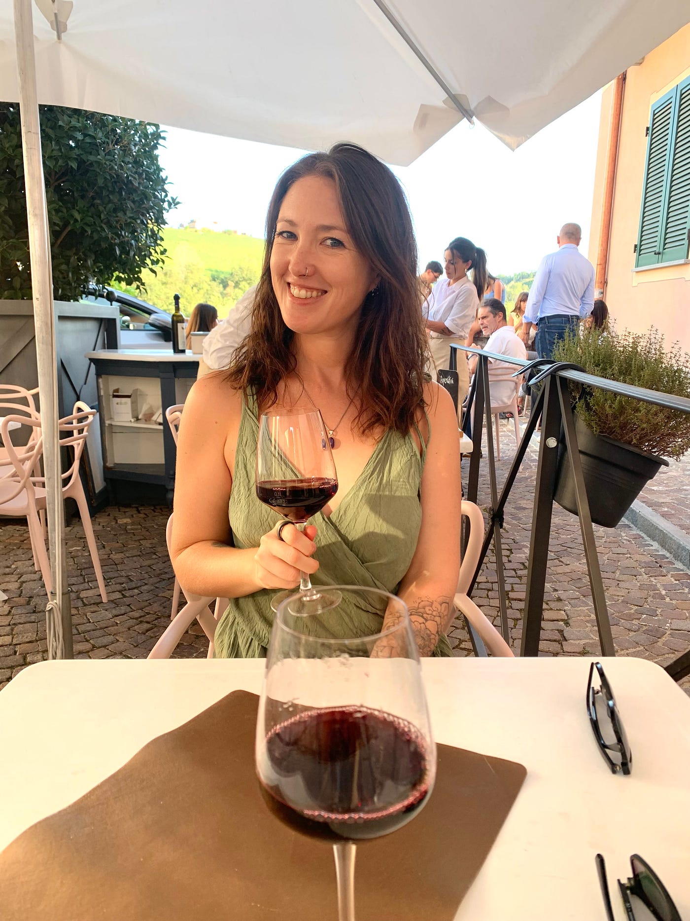 A woman in a green dress smiles and holds a glass of wine to the camera at a bar in Barolo