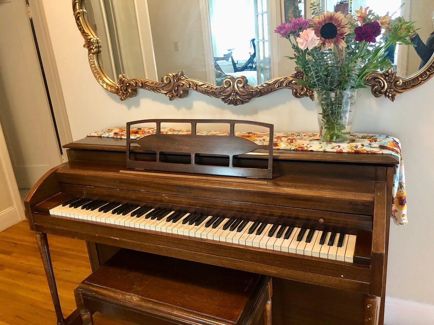 An upright piano made of dark wood under a mirror with an ornate gold frame, with a vase of flowers on top of it.