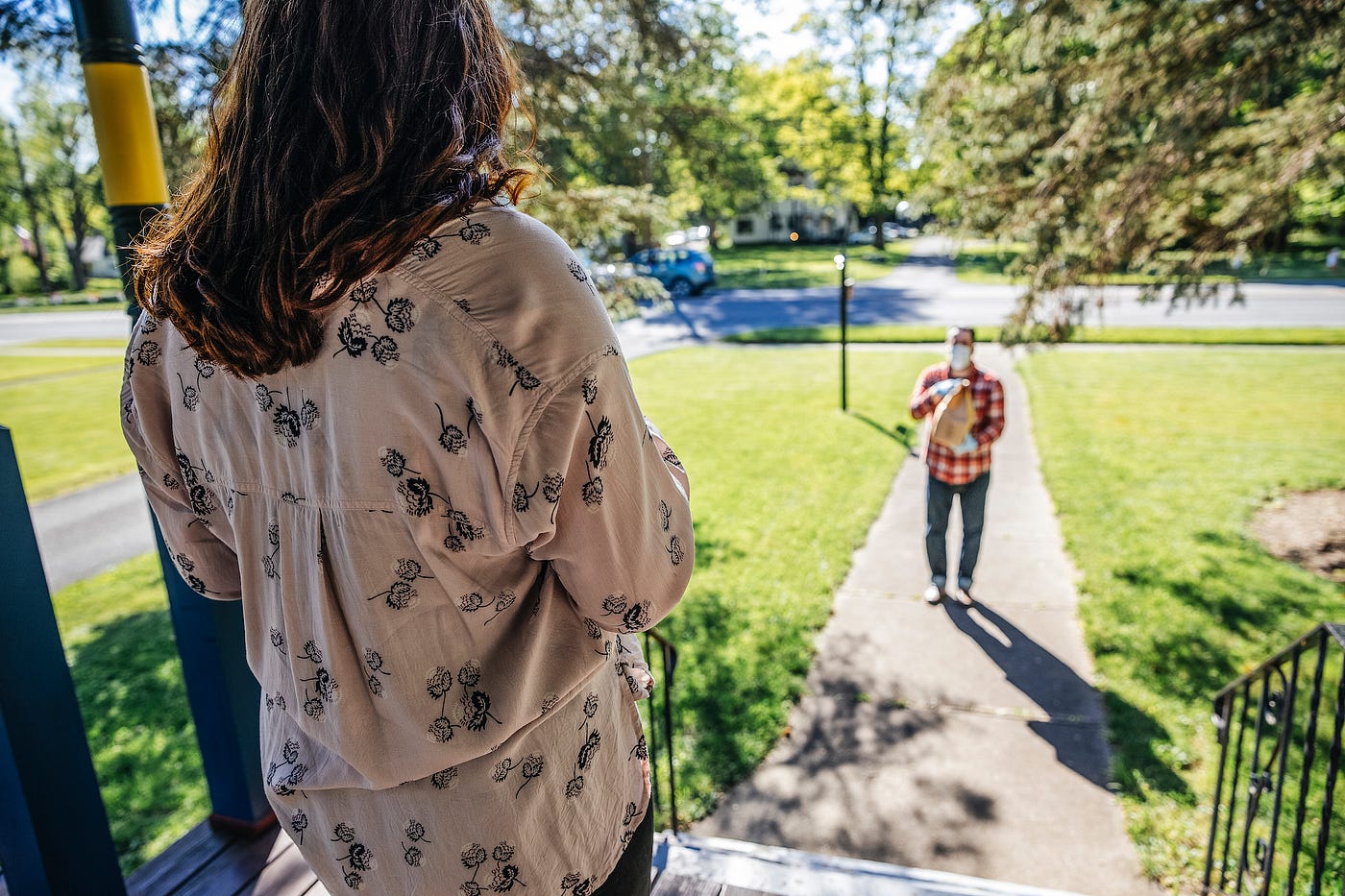 A person on their front porch, looking out at their neighborhood; on their front walk stands a person delivering a bag.