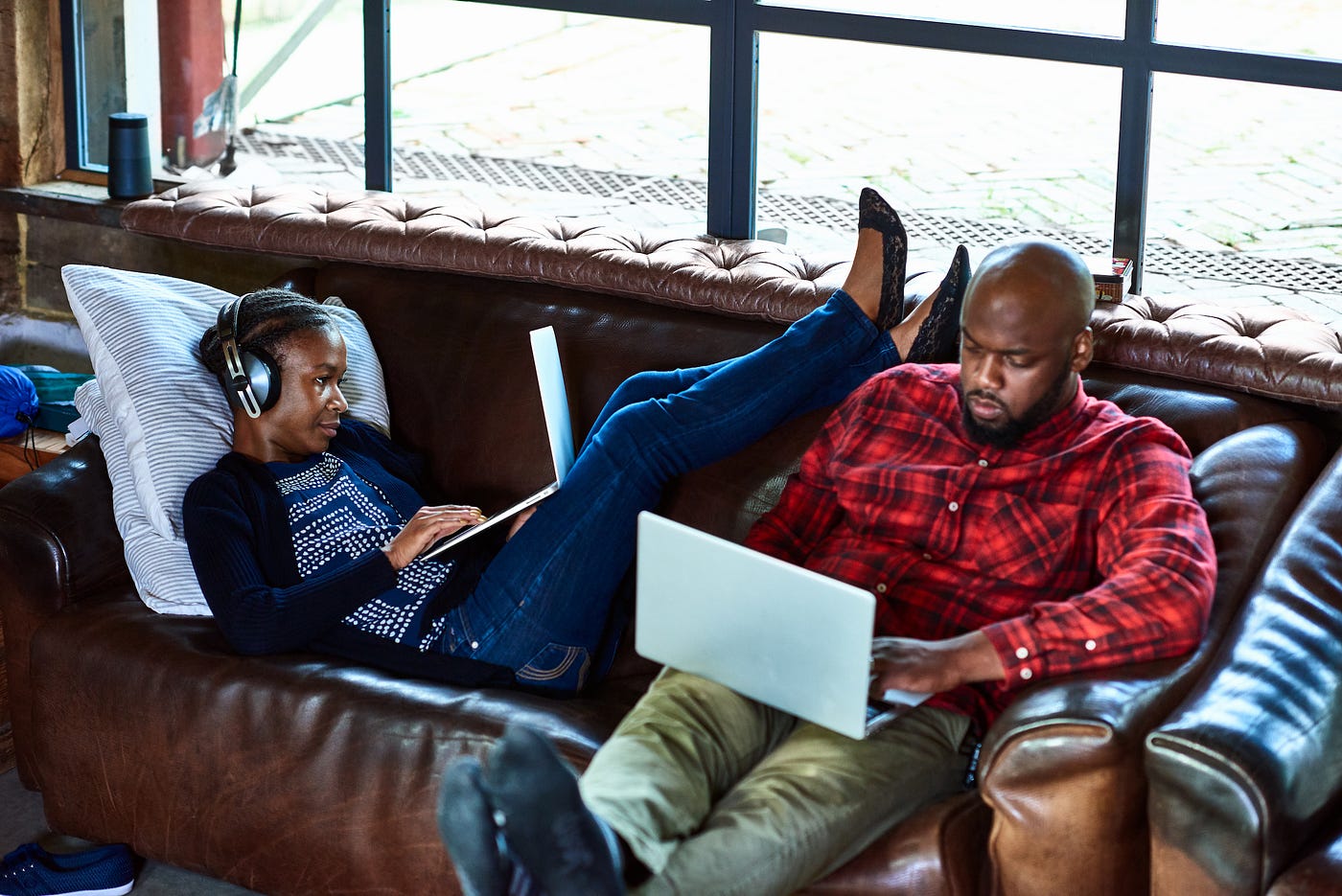 2 people sitting on a sofa working on laptops. One’s reclined, resting their feet on the shoulder of the other, who’s upright