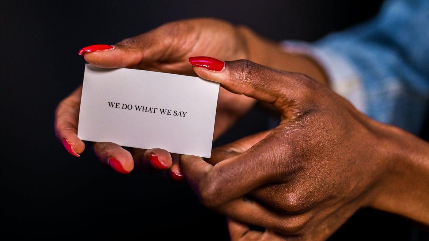 Close-up of a person’s hands with red nail polish holding a white card with the text ‘WE DO WHAT WE SAY’ written on it, against a dark background.