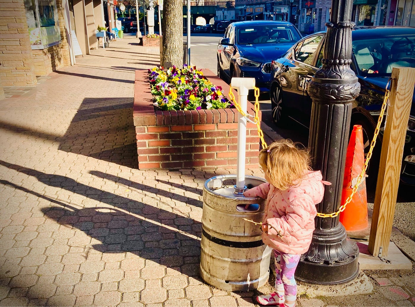 분홍색 코트를 입은 덥수룩한 머리의 아이가 벽돌 보도 위에서 검은색 가로등 옆에 놓인 금속 통을 가지고 놀고 있는 모습이 담긴 사진입니다.<BR> 배경에는 화려한 꽃이 핀 벽돌 화분이 있습니다.<BR>