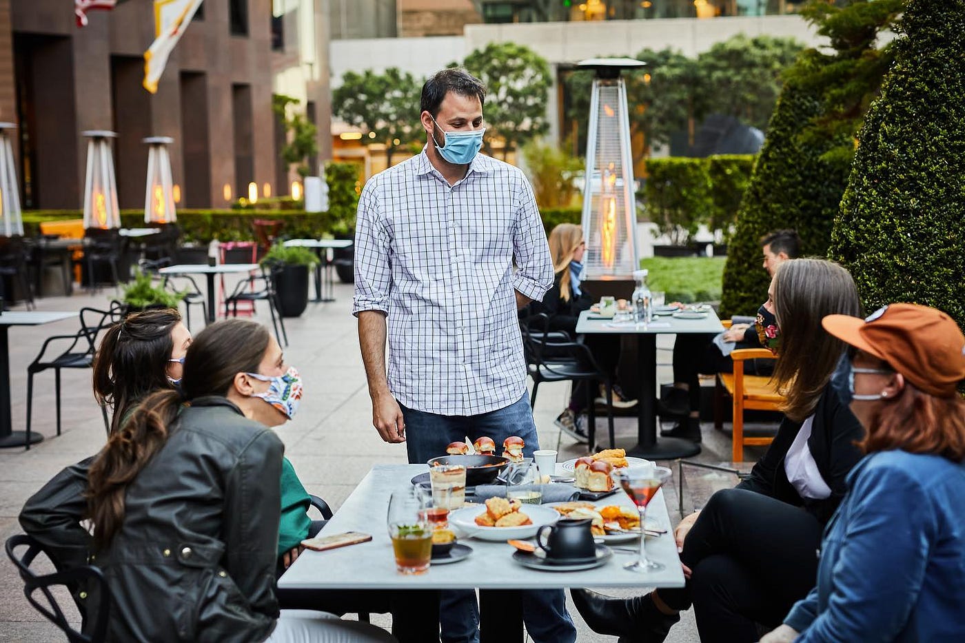 A person standing by a table with four people sitting at it on an outdoor patio. All are wearing face masks.