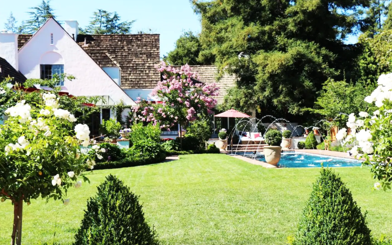 A grassy yard with a rectangular pool with water fountains next to a white cottage.