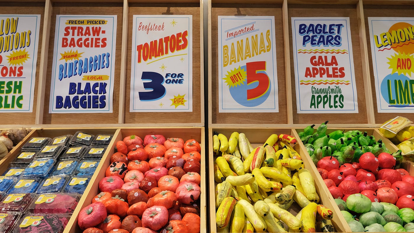 A typical supermarket vista — or is it? Wood crating holds ordinary produce: bananas, berries, apples, and tomatoes. Signage above includes pricing and declaratives such as “Fresh!” But the signs also include text like “baglet pears” and “bluebaggies.” Upon closer inspection, the produce is entirely composed of colorful, single-use plastic bags.