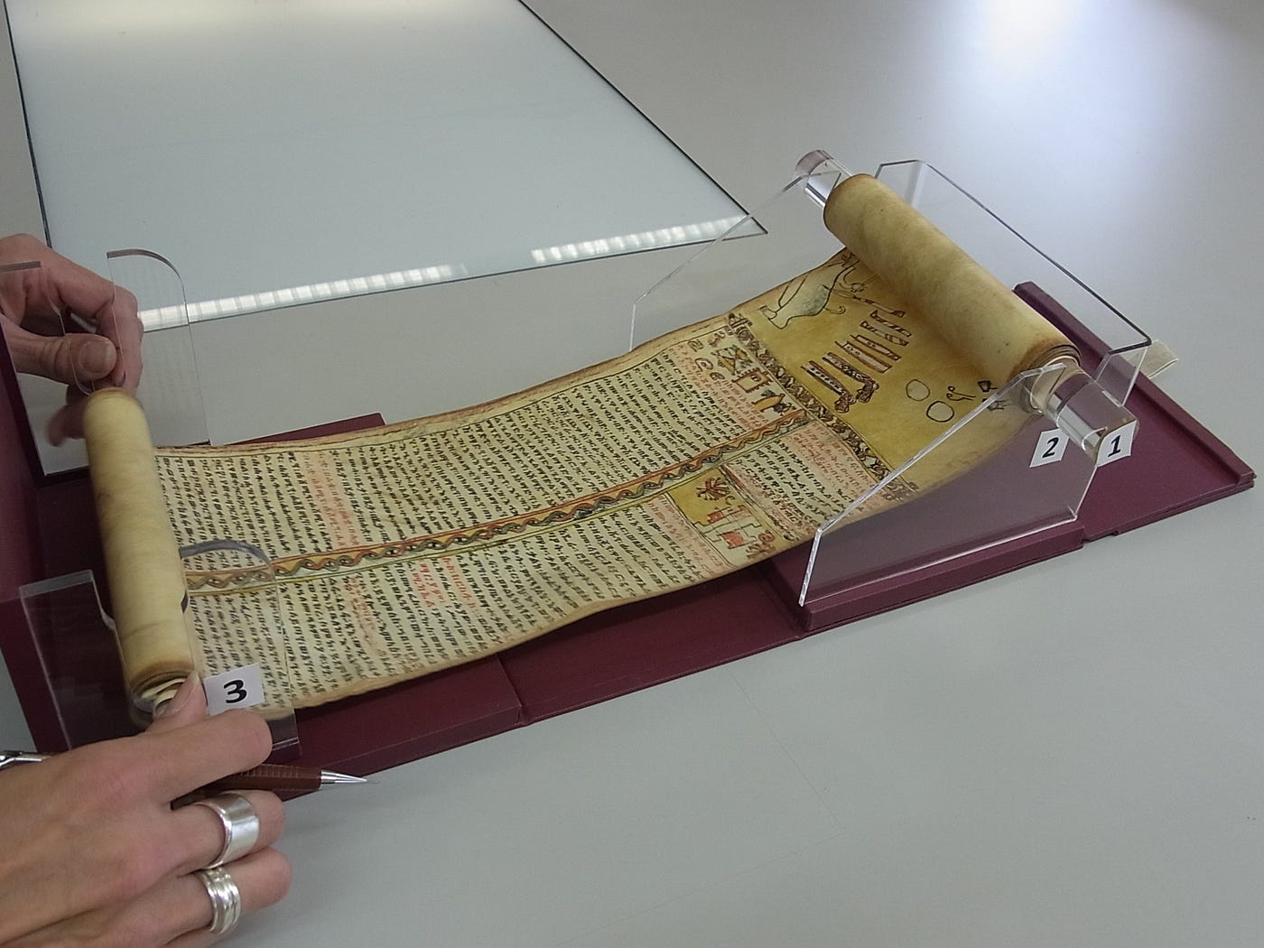 Ethiopian scroll being displayed on Wellcome’s custom-made scroll holder, made of clear plastic. Two hands are unravelling the scroll.