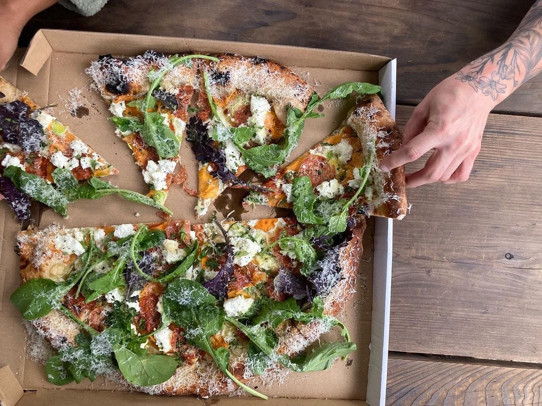 Overhead shot of a hand taking a slice of thin-crust pizza topped with greens out of its box.
