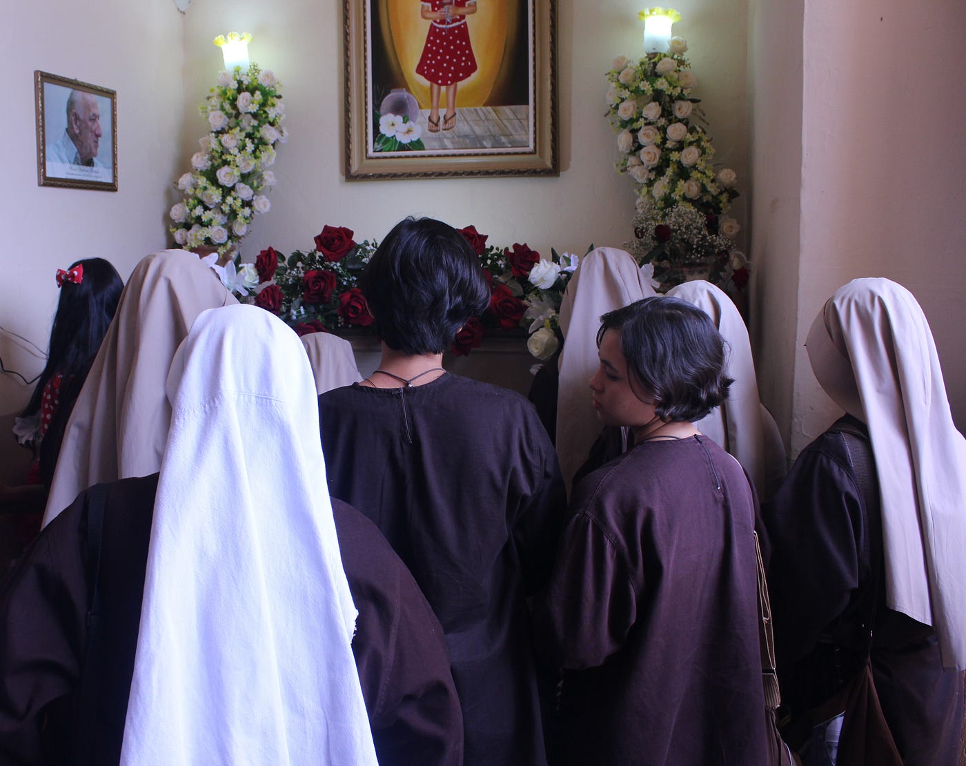 Freiras observam quadro da menina Benigna na Igreja Matriz. As cores de suas roupas e véus destoam do vermelho rubro de Benigna. A fé, no entanto, continua a mesma.