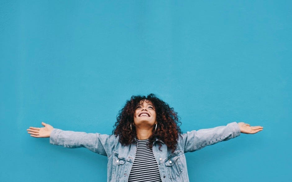 A young woman with dark curly hair in a jean coat against a blue background stands with her arms spread out to each side. She is looking toward the sky and smiling in a celebratory pose.