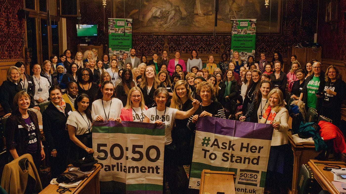 A large room in Parliament, full of women smiling with banners reading 50:50 Parliament.