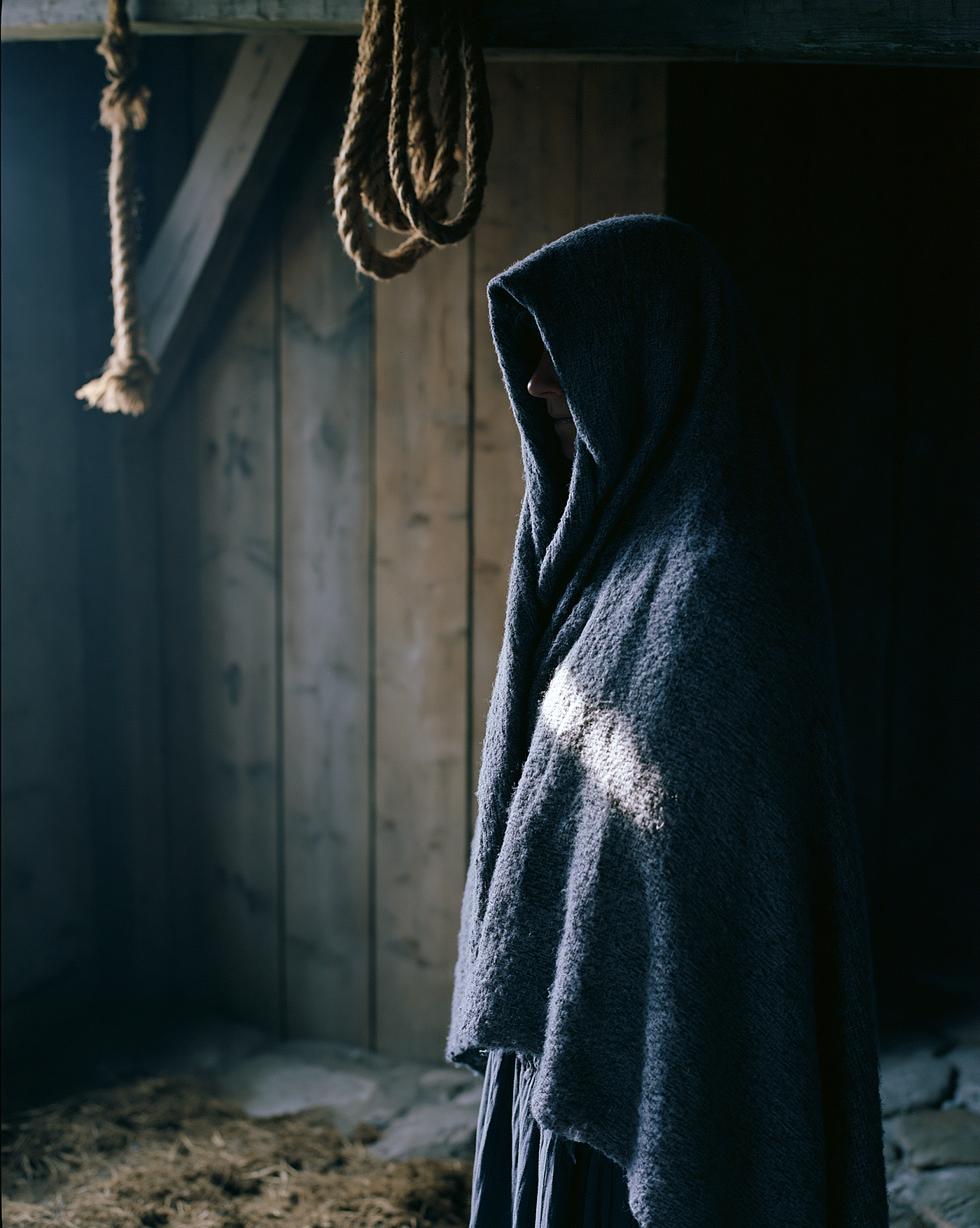 Prompt: documentary photograph of a middle-aged woman standing motionless in a dim unheated barn on the island of Fanø, her face and head completely wrapped in a coarse hand-woven strude veil leaving only her silhouette visible, photographed in the spirit of Trine Søndergaard, shot on Mamiya 7 II with 80mm f/4 using Kodak Ektar 100, single crack of grey overcast daylight entering through a gap in the wooden planks casting a narrow blade of cold light across the veil texture, desaturated palette of weathered timber brown and damp grey wool and blue-cold skin undertones, rough hemp rope hanging from ceiling beams and dried mud on stone floor, figure positioned off-center against the darkest wall, the veil texture sharper than the background, fine controlled grain with no highlight bloom and deep unlifted shadows — ar 4:5 — raw — stylize 80 — hd — v 8.1