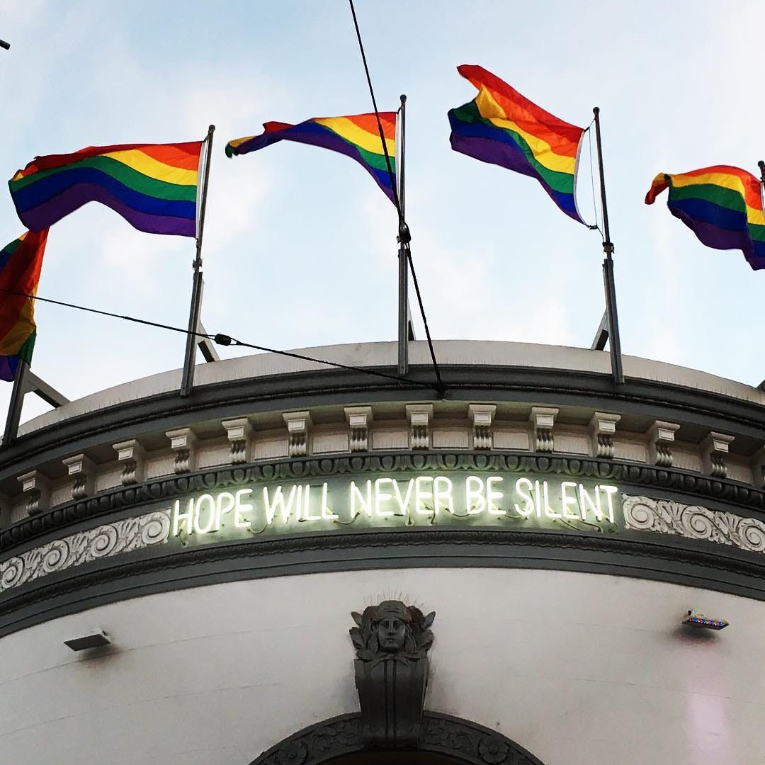 A rounded building with “Hope will never be silent” in neon lights near the roof, which has 5 rainbow flags on it.