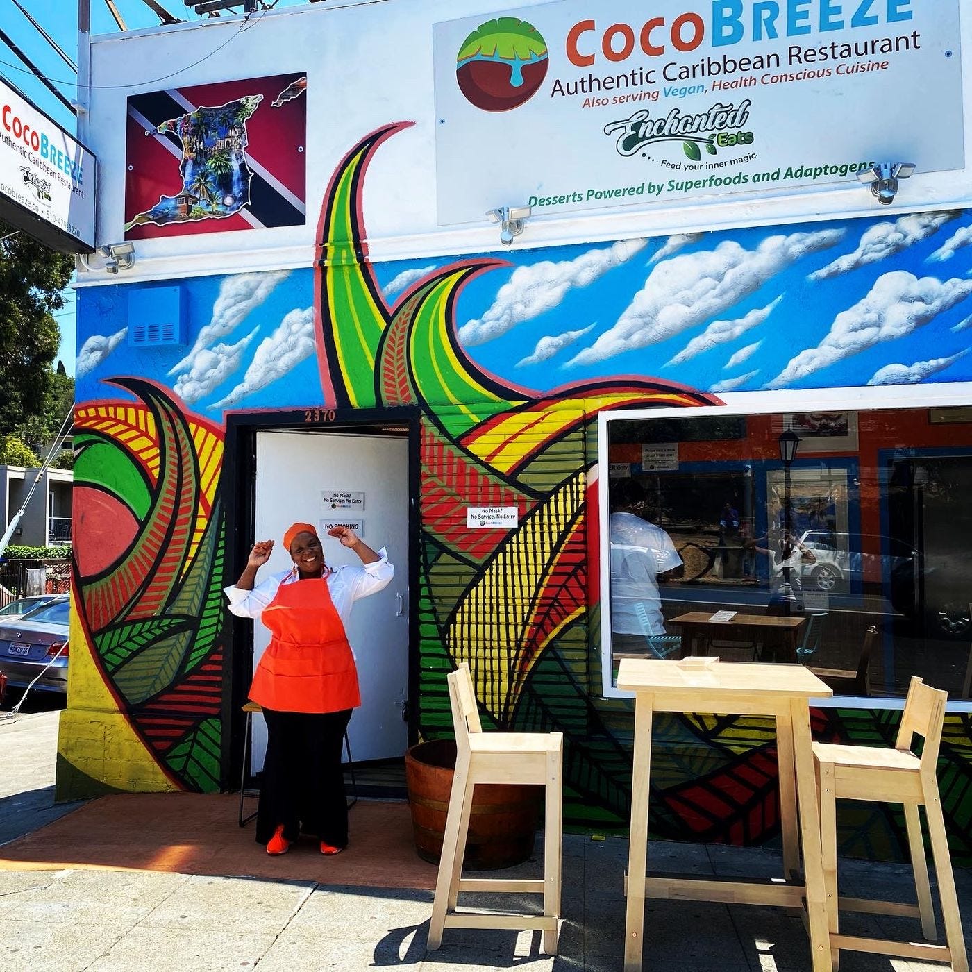 A person wearing a red apron smiles, arms raised, at Coco Breeze’s front door.