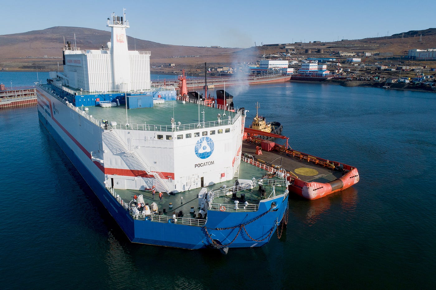 A large white-and-blue ship with equipment on its decks, docked in a body of water near the shore.