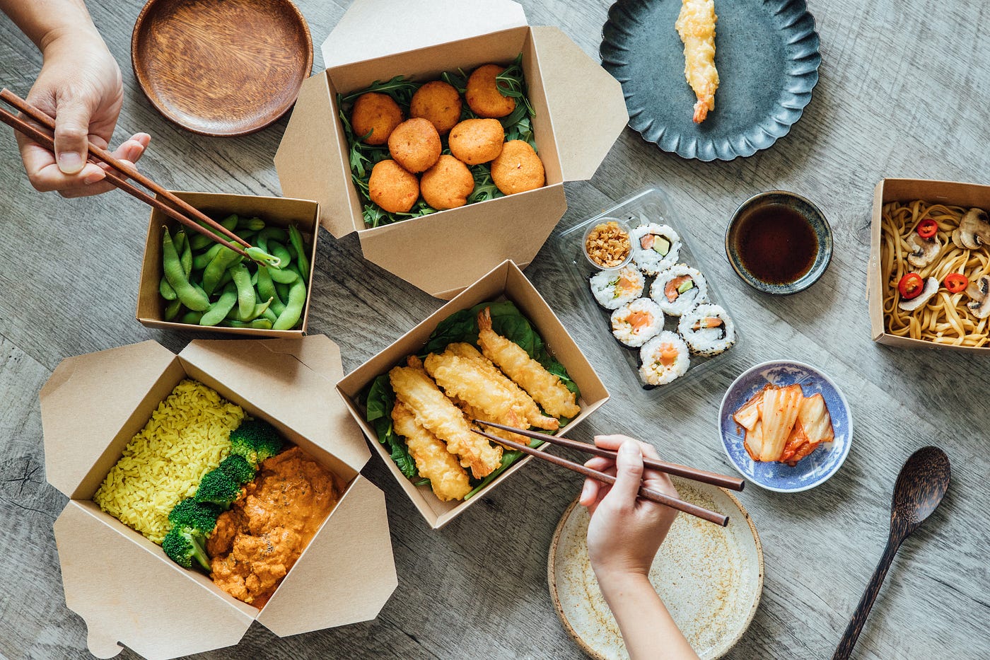 Overhead view of a variety of Asian carryout dishes on a table with people’s hands holding chopsticks.