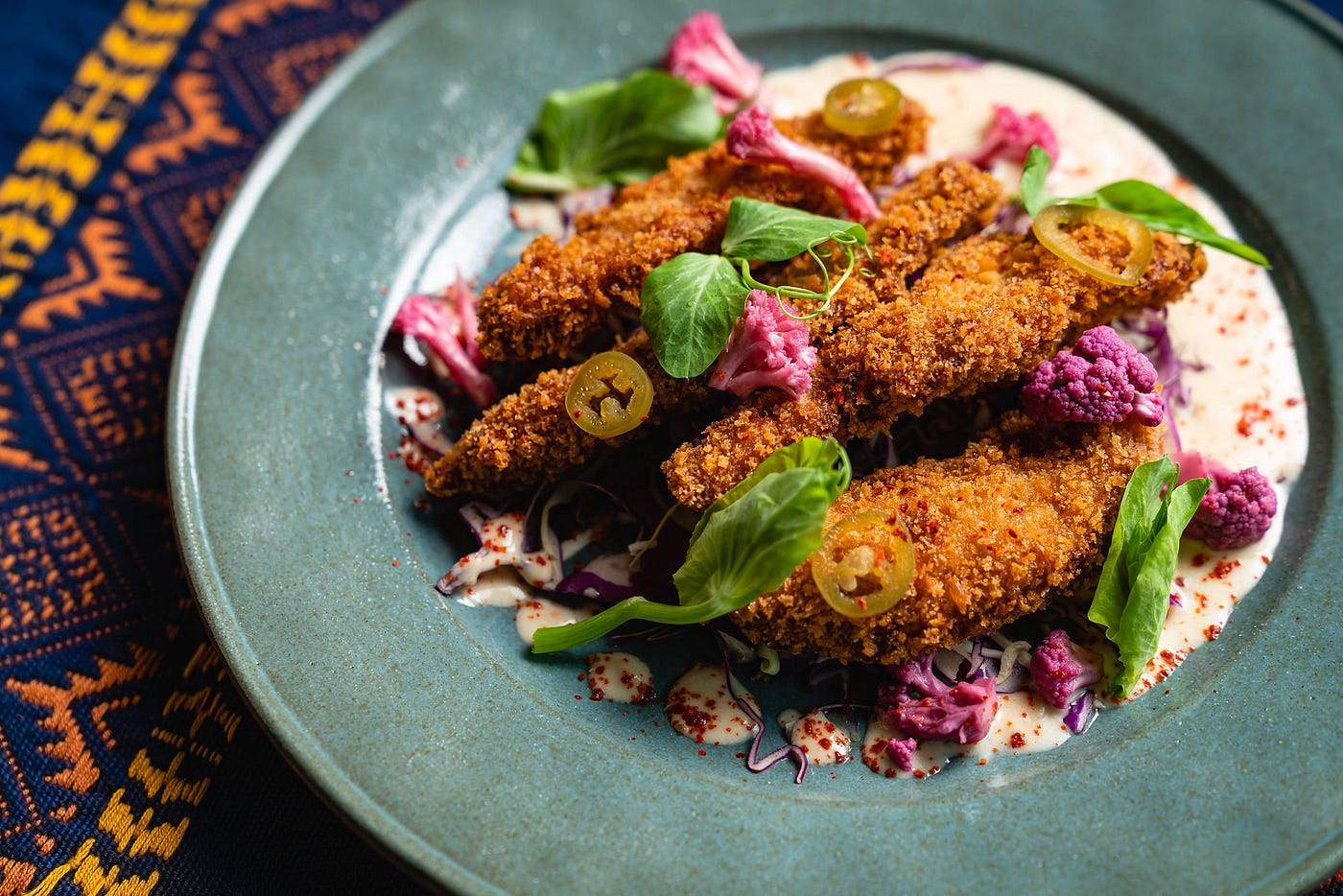 Stripes of fried and breaded pork loin in coconut sauce in a bowl on top of a tablecloth with a geometric pattern.