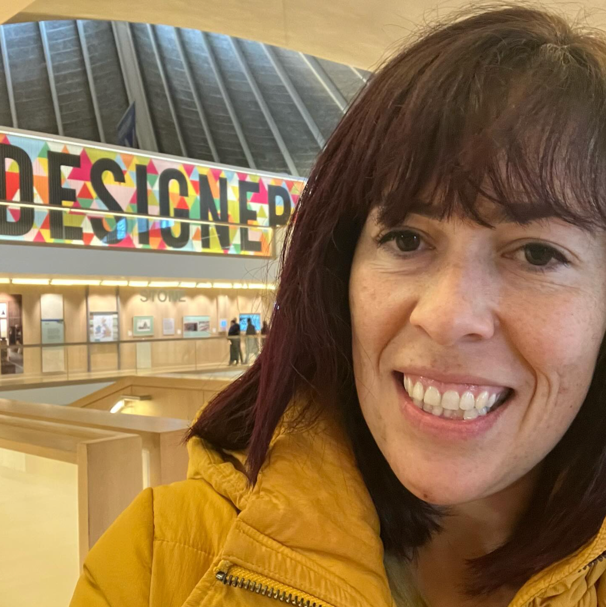 A woman with pink brown hair, fair skin with freckles and dark eyes smiles in front of a wall that say 'Designer'