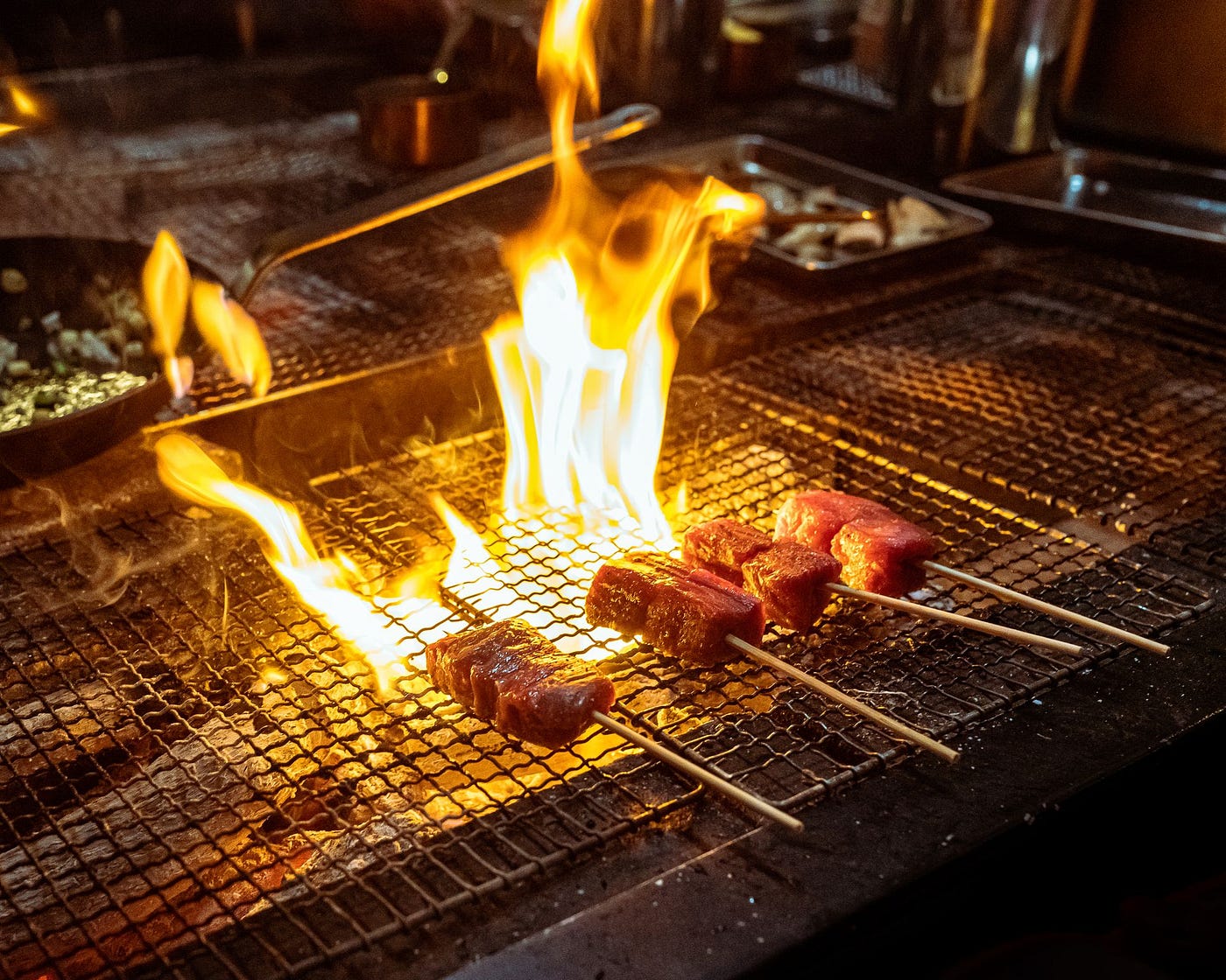 4 meat skewers on a wire rack over a grill, lapped with tall flames.