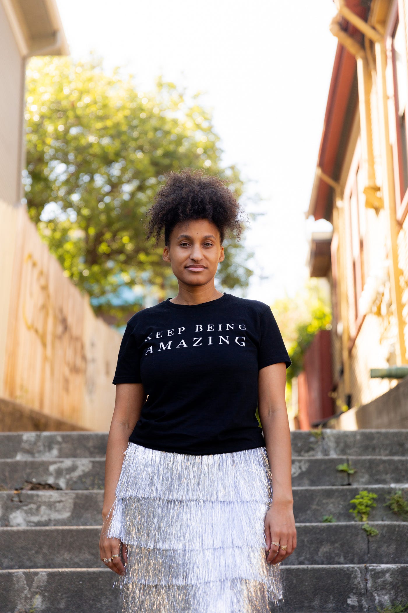 Akilah standing in a sunny alley wearing a black T-shirt that reads, “Keep being amazing.”