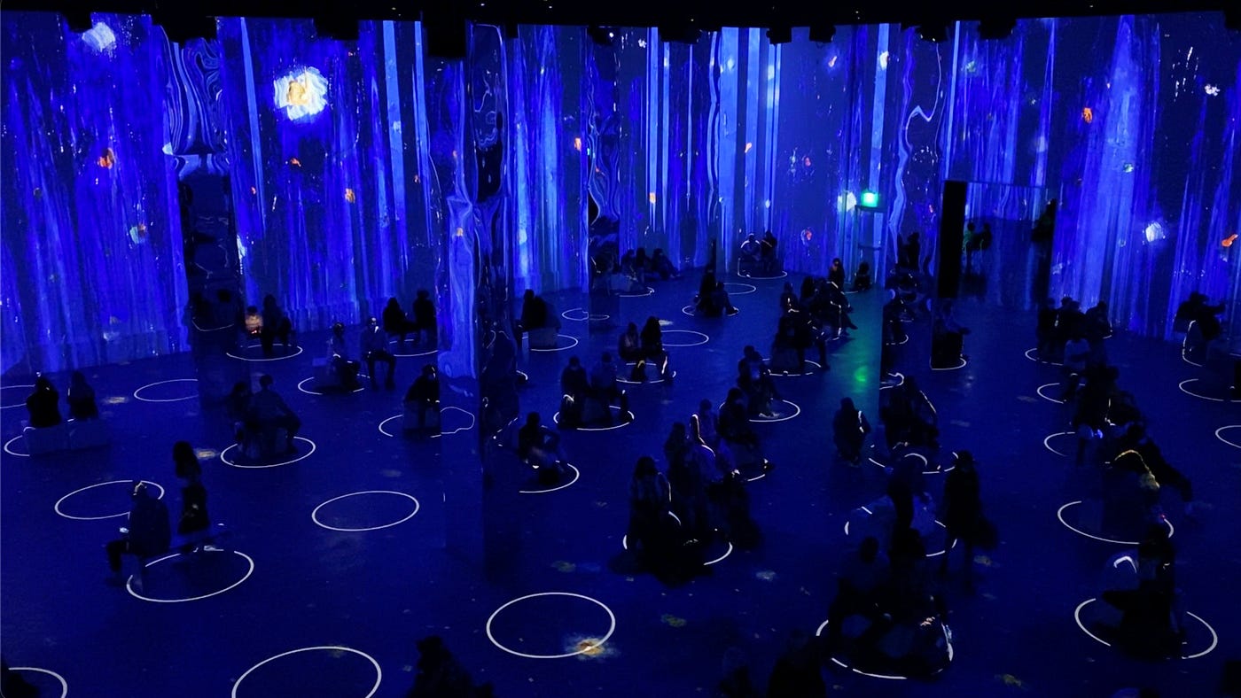 The image is of a dark, cavernous room, which is viewed at an angle, from above. The entire room is bathed in a deep marine blue, with streaks of white along the walls. Looking down, there are circles of white evenly spaced apart. Silhouettes of people in small groups, mostly pairs, fill each circle’s outline.