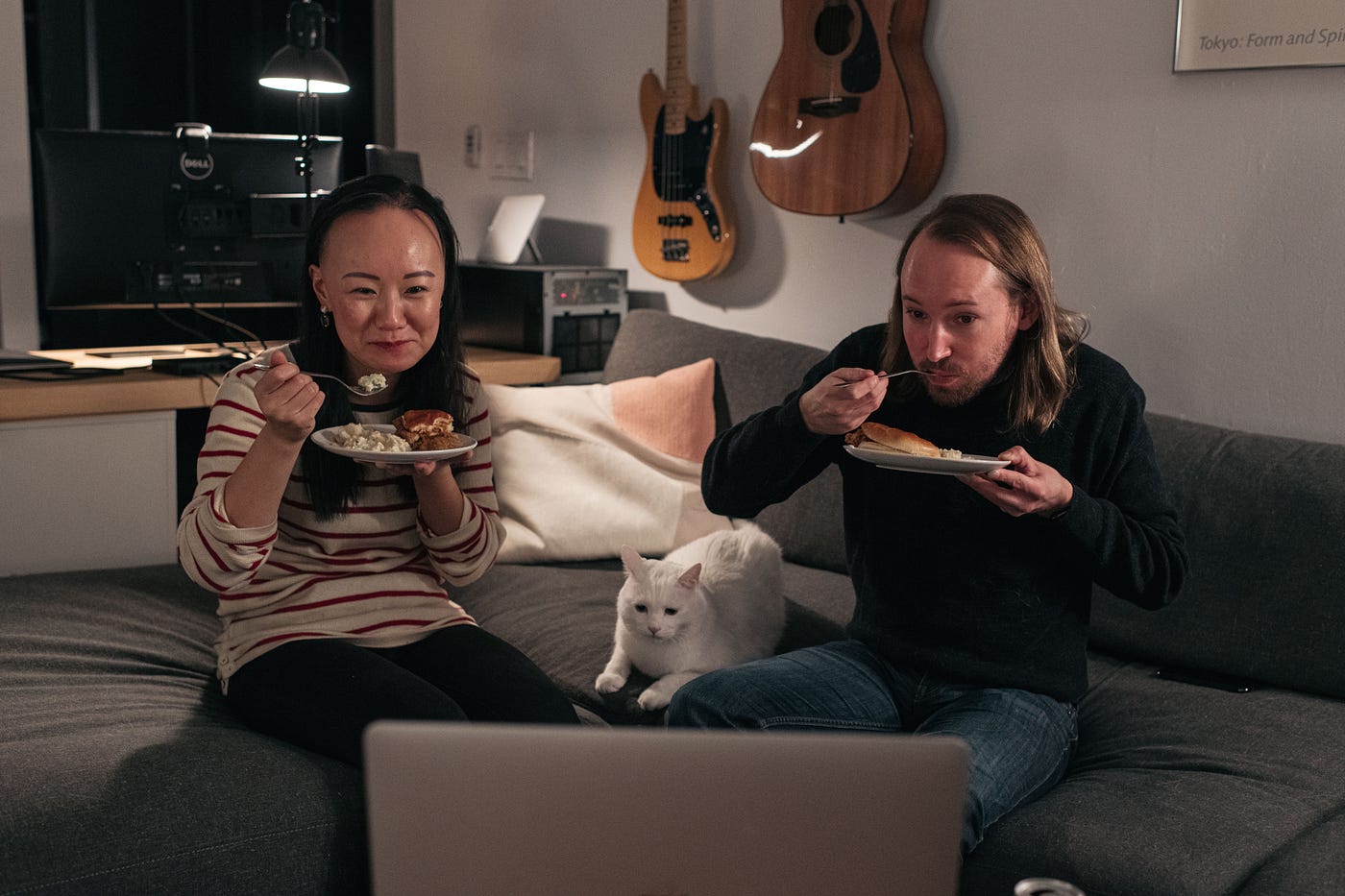 A couple + a white cat sitting on couch in a living room with guitars on the wall, eating and watching a laptop.