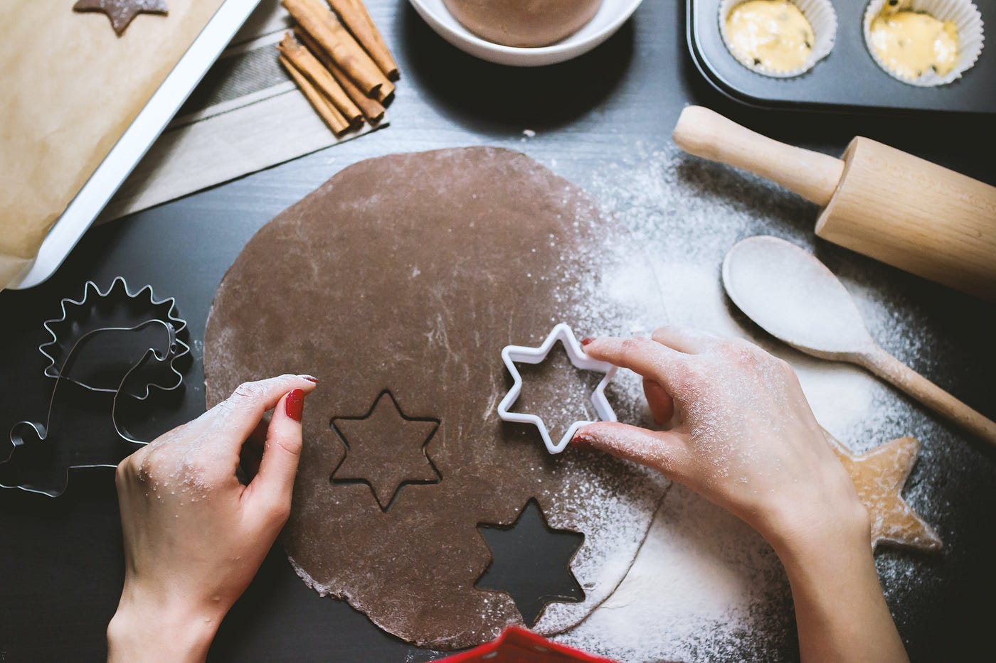 A person’s hands pressing star-shaped cookie cutters into chocolate cookie dough, with a rolling pin, scattered flour, and additional baking utensils in the background.