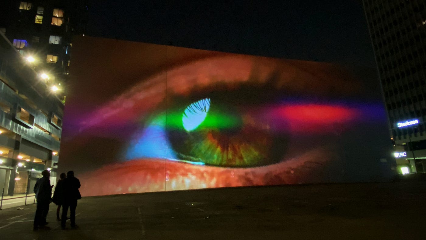 An urban parking lot at night. The image of a human eye is projected onto the entire, windowless wall of a building. Silhouetted in the foreground are a few people, looking tiny in comparison to the scale of the projected eye.