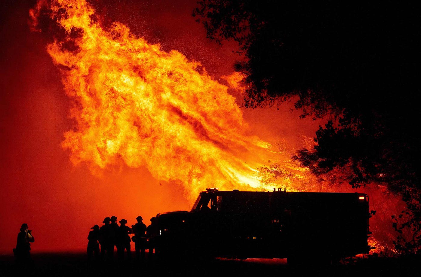 Firefighters standing next to their fire truck, silhouetted by flames that are several times taller than the truck.