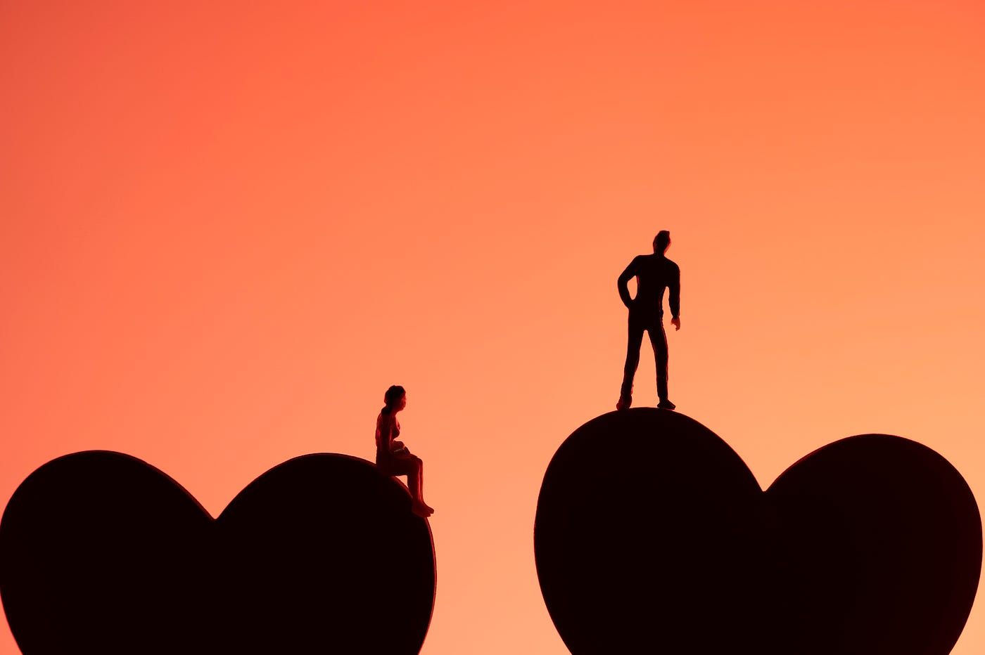 Silhouettes: A person sits on a large heart are the bottom lefthand corner of the photo; another person stands on another large heart in the bottom righthand corner. The black silhouettes are set off by a pinkish/salmon background.