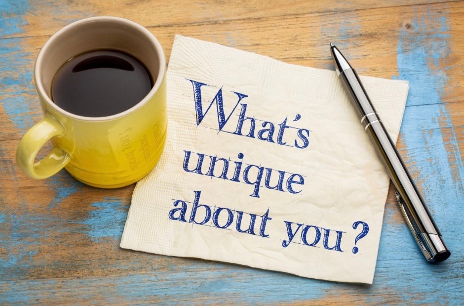 A wooden table with blotches of blue paint. There is a yellow coffee cup, silver oen and a napkin that says: WHAT’S UNIQUE ABOUT YOU?