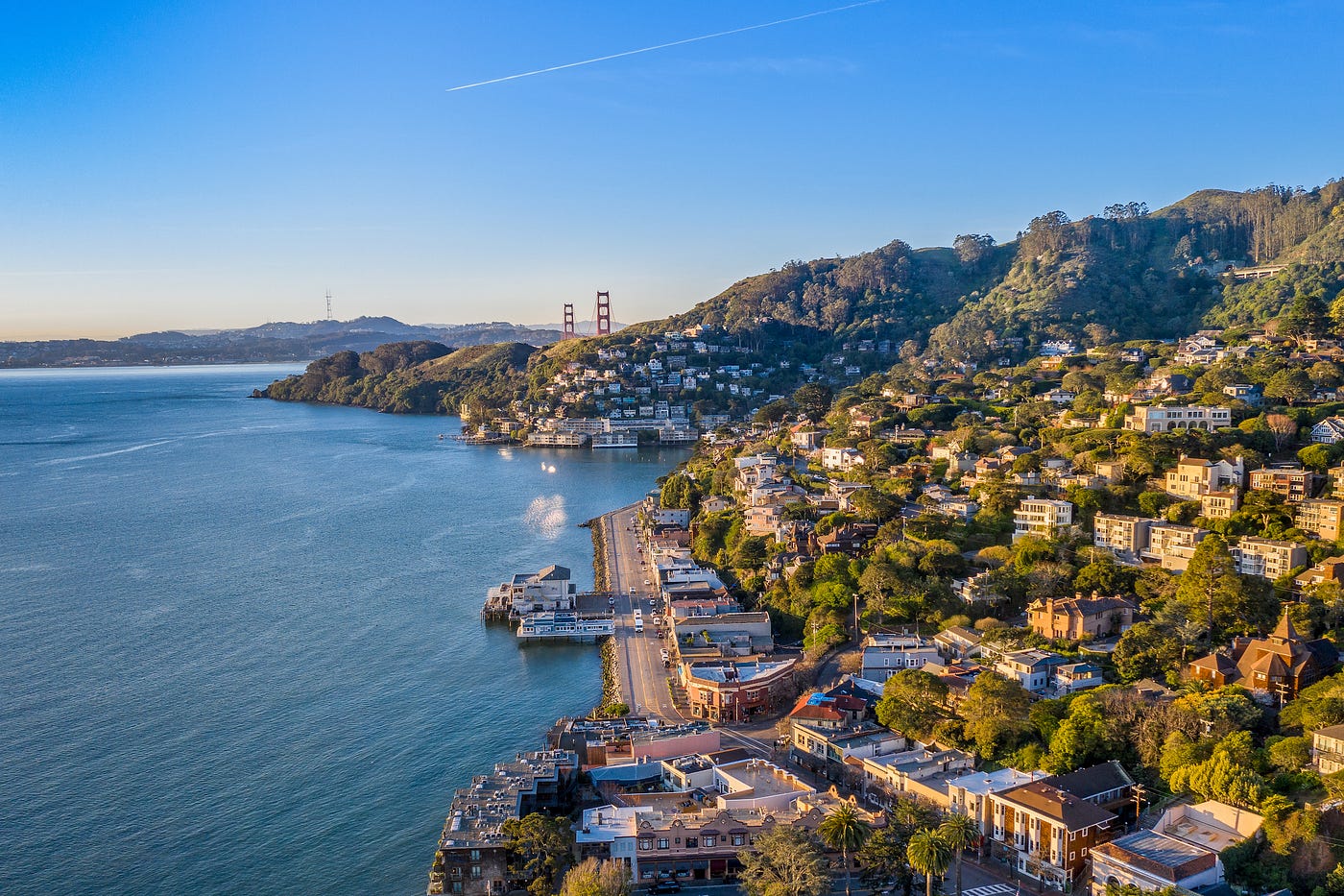 Aerial view of the coast near Sausalito in Marin County. The Bay is on the left side of the screen and the land on the right. A road hugs the coastline, disappearing behind a hill. Behind more hills, far in the distance, is the Golden Gate Bridge.