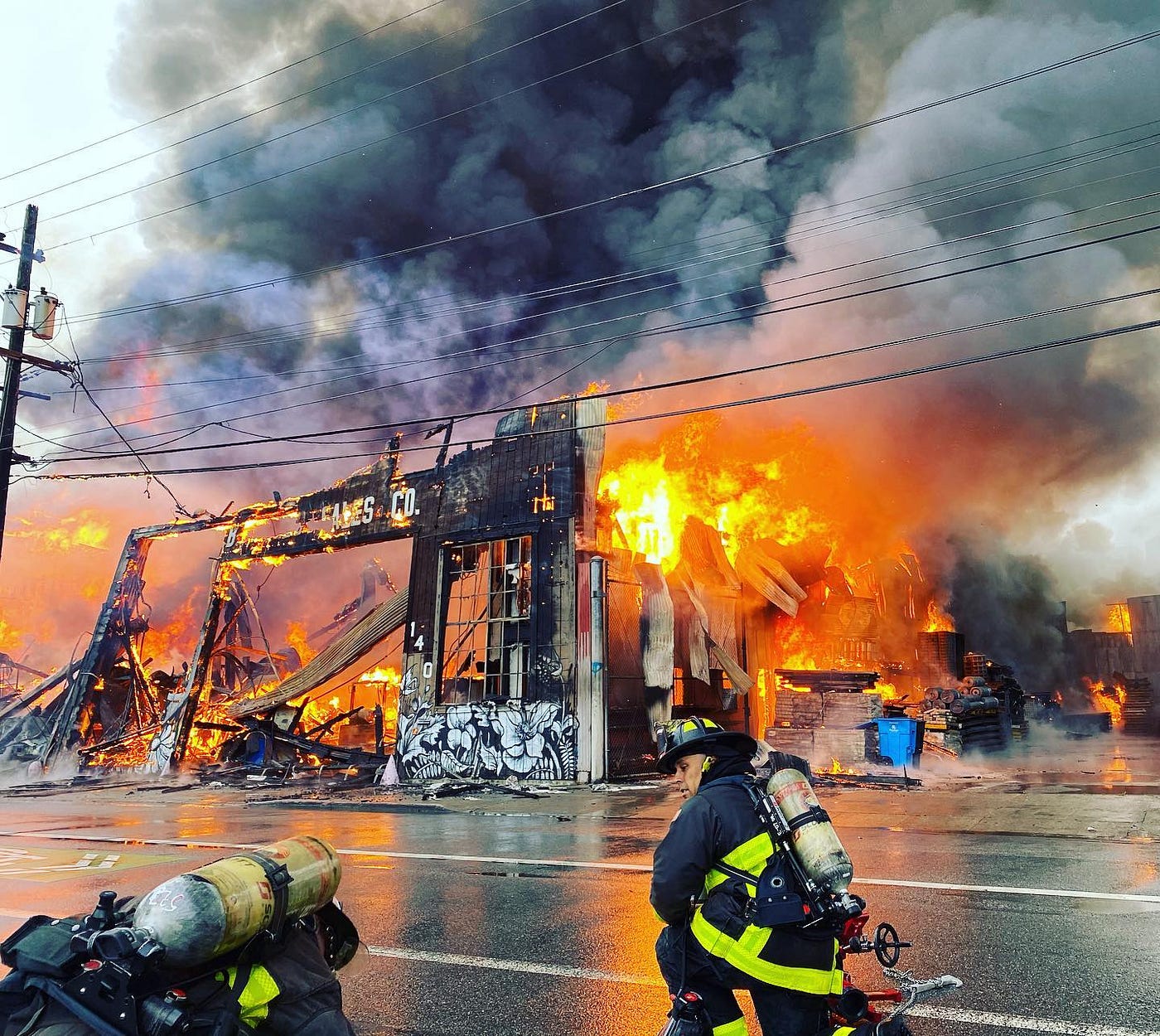 Firefighters standing across the street from a burning building that’s been reduced to the studs.