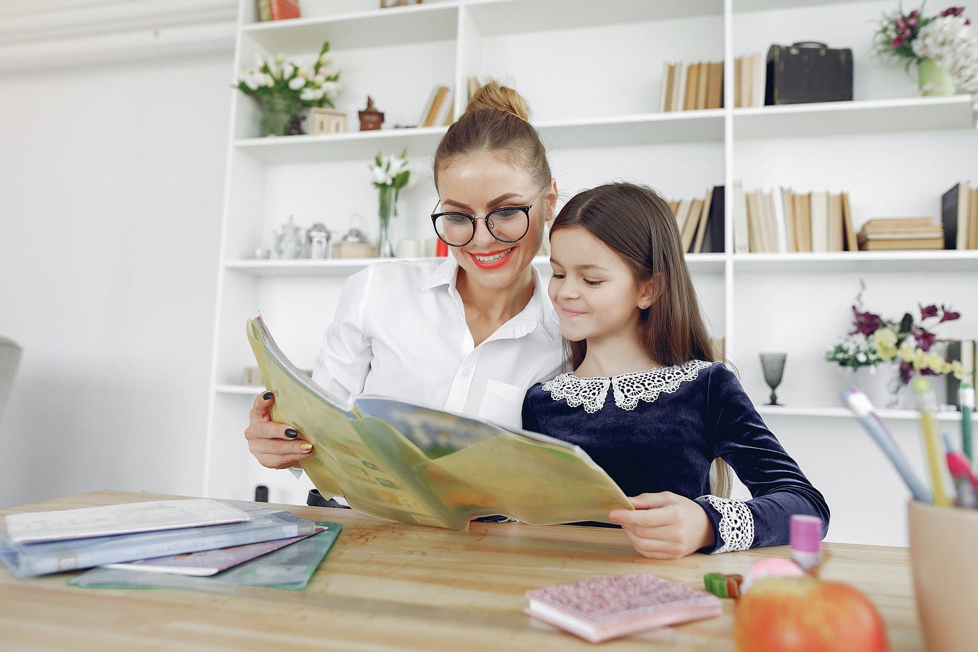 A woman and a young girl are smiling and reading a book together at a desk in a room with bookshelves and a bright, cozy atmosphere.