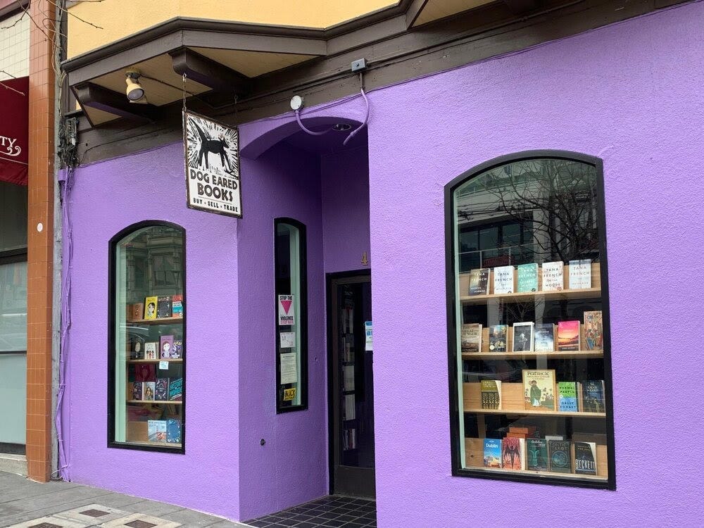 Exterior of a shop with bright purple stucco and windows showing bookcases of books on either side of its front door.
