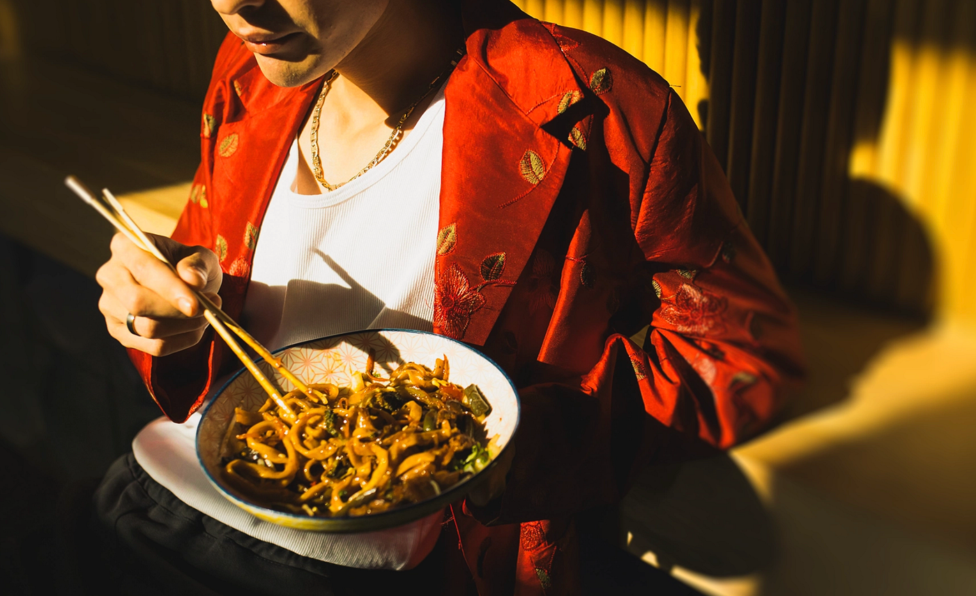 Close-up shot of person in red floral jacket eating stir-fried noodles with chopsticks, highlighting cultural dining and warm dramatic lighting.