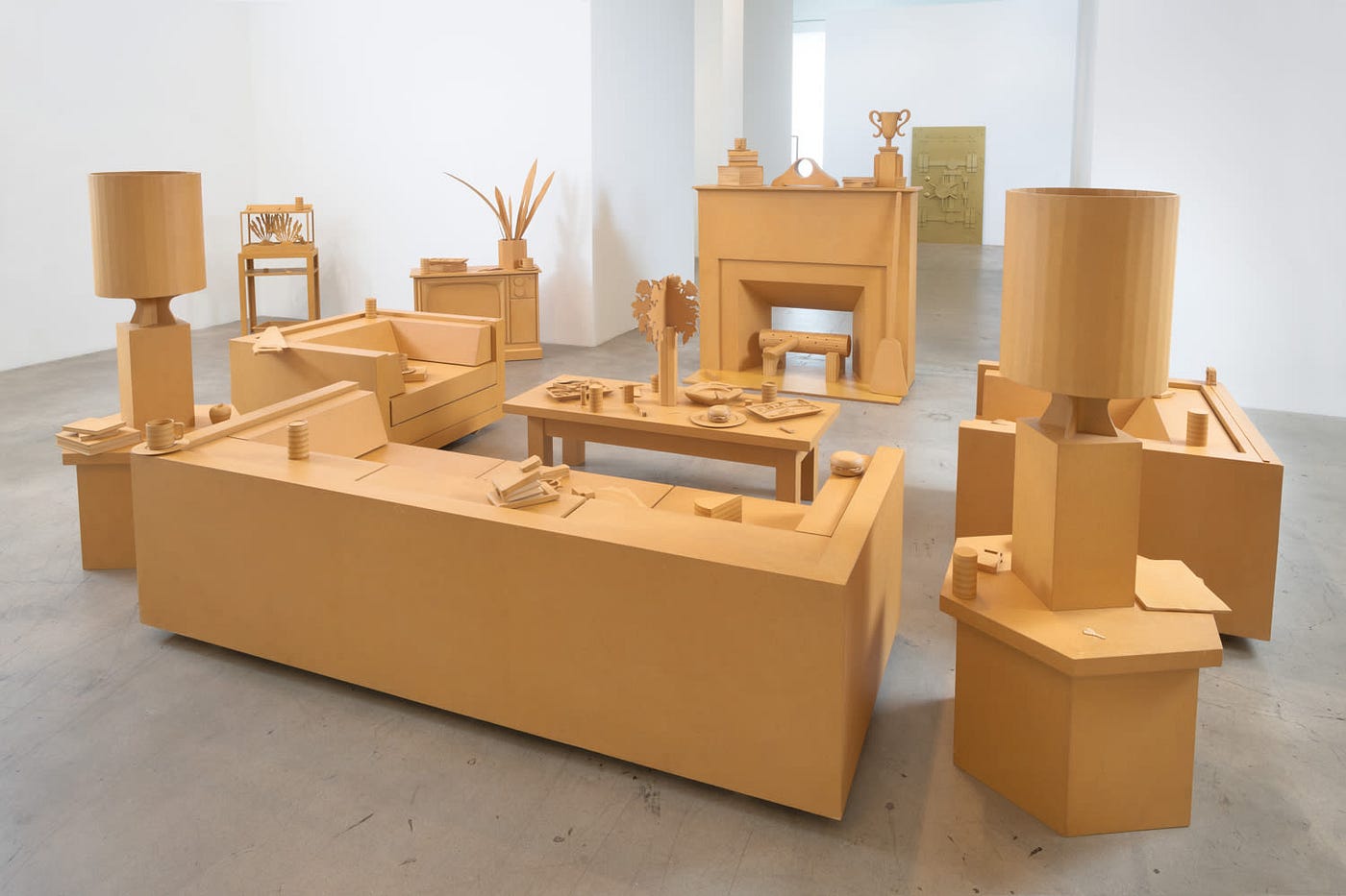 Inside a white gallery space is a freestanding living room made entirely of cardboard, which is a uniform, yellow-brown color. The focal point is a central fireplace, with items on the mantle and logs in its hearth. Directly across from the fireplace is a couch, with armchairs on either side and a coffee table in between. Various objects such as TV dinner trays, with leftover food, are strewn about. Table lamps, an aquarium, and a TV set complete the scene.