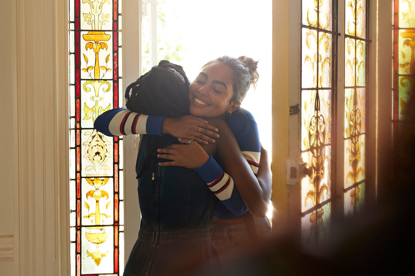 Two female friends embrace in front of an open door with stained glass windows.