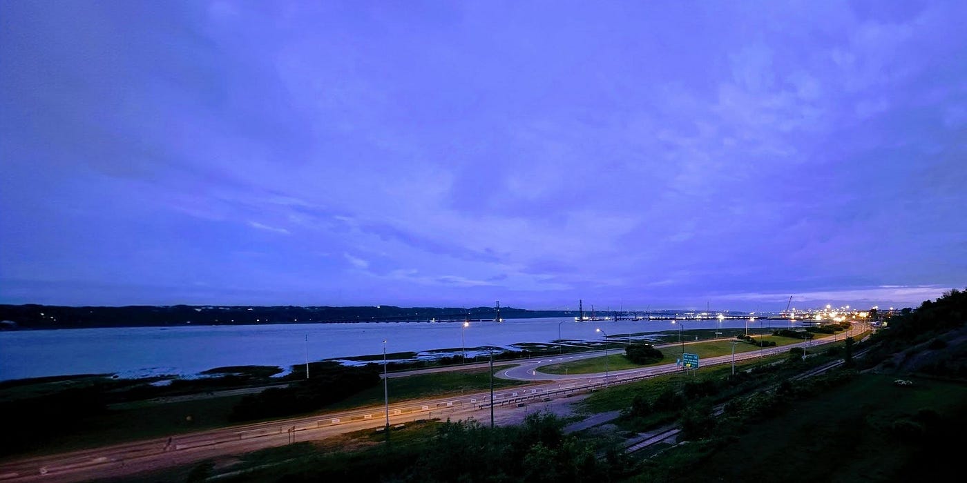 Nocturne Cobalt: The deep indigo sky of the blue light hour hanging heavily over a the St.-Lawrence River. Winding coastal roads lit by warm, amber streetlamps trace the shoreline below, leading the eye toward a distant, illuminated bridge structure on the horizon. (Photography: Francine Fallara)