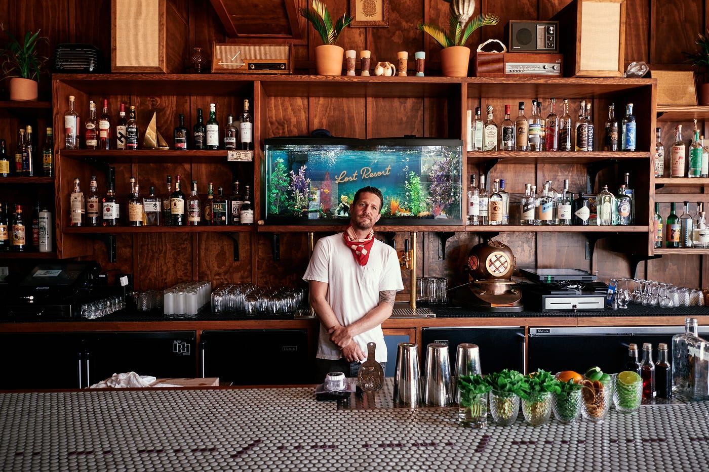A person wearing a white T-shirt and a red bandanna around their neck posing behind a bar.