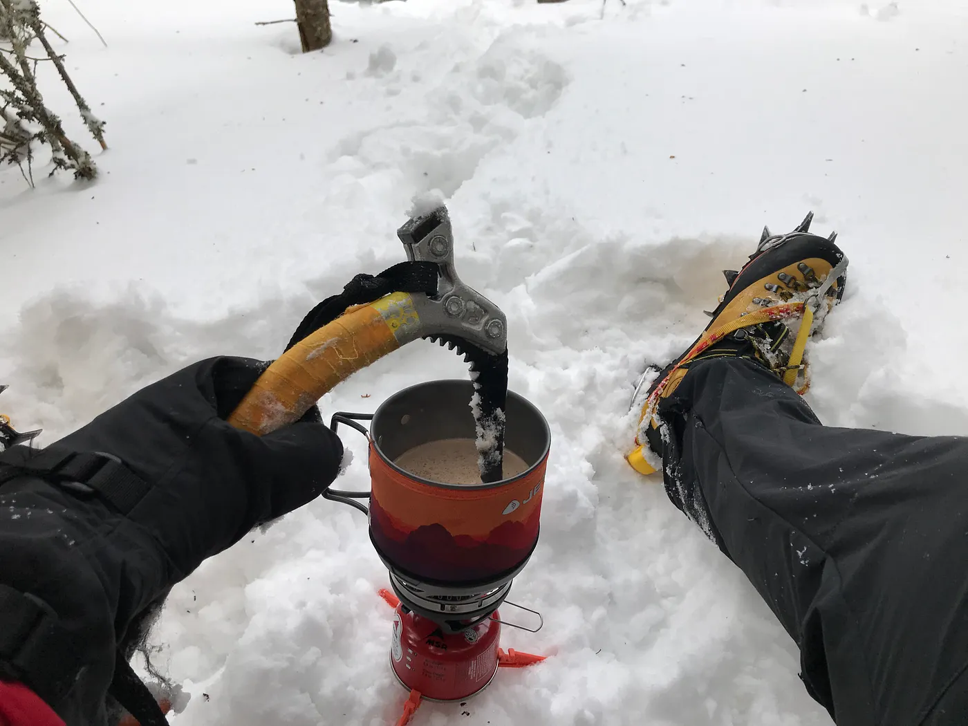 A mountaining stirring a hot drink over small gas stove with an ice climbing tool.